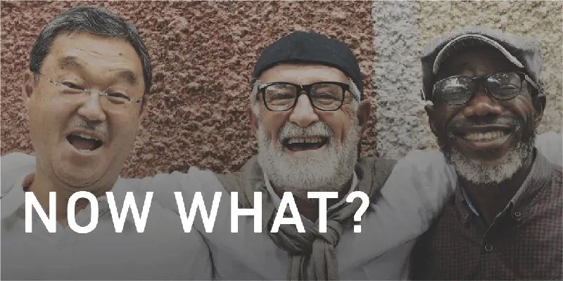 3 older men of different nationalities stand together with arms around each other. All men are smiling and are in their senior years.